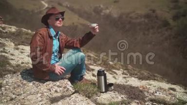 山上坐着一个男人，戴着牛仔帽、皮夹克、蓝色牛仔裤和眼镜。 一个人<strong>喝热水</strong>瓶里的茶。
