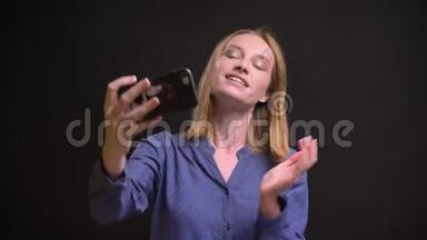白种人金发女人穿着衬衫在黑色背景下使用智能手机制作自拍照片的肖像。