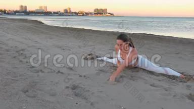 日出时，坐在沙滩上做瑜伽的年轻女子在沙滩上劈叉。