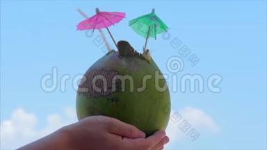 年轻的双手女人在热带海滩上的美丽的云彩背景中抱着椰子。 暑假的概念