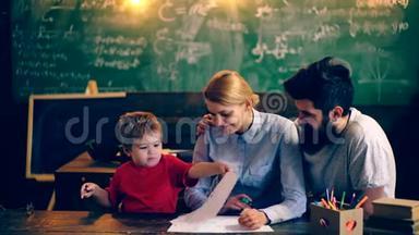 男孩和他的父母在学校教室里画黑板的背景。 父母教他们的儿子。 教师