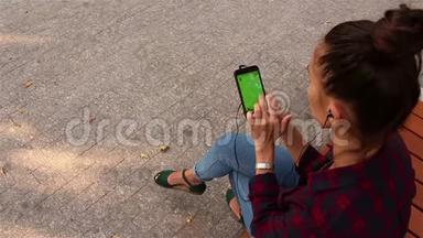 年轻漂亮的女孩坐在公园的长凳上，在智能手机上浏览社交网络。