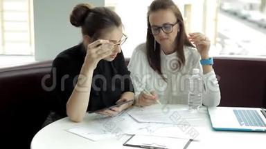 两个戴眼镜的女商人坐在一起讨论生意，使用平板电脑和笔记本电脑，绘制商业发展图表