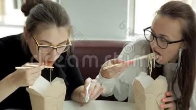 两个戴着眼镜的年轻商务女孩坐在笔记本电脑的咖啡馆里，吃着中国面条，看着笔记本电脑
