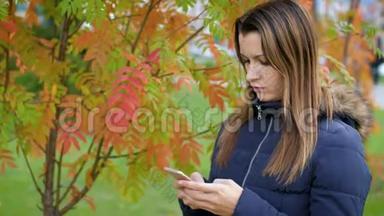 一个年轻的女人<strong>拿</strong>着一部<strong>手机</strong>站在鲁文脸红的灌木丛前。 女孩用智能<strong>手机</strong>站在