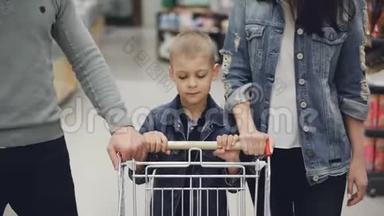 可爱的孩子在<strong>食品店</strong>里推着购物车，他慈爱的父母在帮助他。 儿童、青年家庭