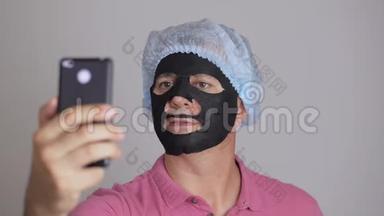 一个男人，把一个恢复活力的黑布化妆品面膜敷在脸上，用智能手机自拍