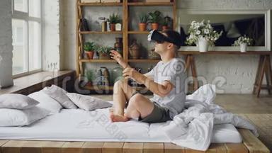 兴奋的年轻人正在使用增强现实眼镜移动双手，同时坐在轻型公寓的双人床上。 现代