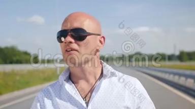一个戴太阳镜的秃头男人在路上唱歌和散步的肖像。 中弹。