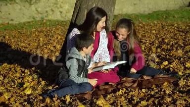 妈妈和孩子们坐在树下看书。 秋天森林里的妈妈和孩子们.. 他们很开心。