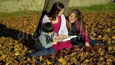 妈妈和孩子们坐在树下看书。 秋天森林里的妈妈和孩子们.. 他们很开心。