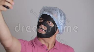 一个男人，把一个恢复活力的黑布化妆品面膜敷在脸上，用智能手机自拍