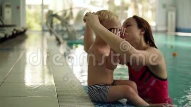 一个小游泳运动员准备游泳。 这个男孩坐在游泳池的边缘，他妈妈戴上眼镜游泳