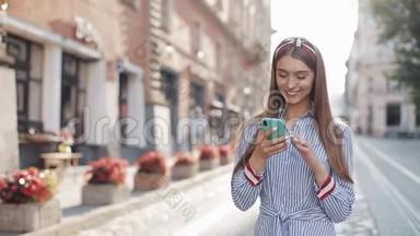 微笑的年轻女子穿着蓝色和白色条纹连衣裙，用智能手机在老街上<strong>走来走</strong>去