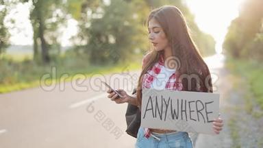年轻漂亮的女人<strong>搭车</strong>站在路上，拿着任何地方的标志，用智能手机。 少女