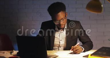 年轻的黑人商人带着笔记本电脑和文件在夜间办公室工作。 他在文件上<strong>签</strong>字。 商务