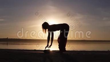 一个身材匀称的女人在水边的剪影展开瑜伽垫，准备做瑜伽或普拉提。 日落海滩