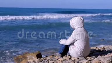 穿着白色羽绒服和风帽的女人坐在岸边，看着<strong>汹涌</strong>的<strong>海浪</strong>拍打着石头