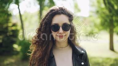 戴着太阳镜和时髦皮夹克的迷人女人站在城市公园里看镜头和微笑的肖像