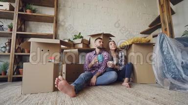 可爱的一对夫妇在搬迁后坐在新公寓的地板上交谈、亲吻和抱着杯子。 许多箱子