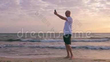 日出时，在波浪海附近的海滩上，男人在智能手机上自拍