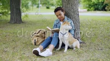 快乐的混血女孩狗主人正在看书，并抚摸她可爱的<strong>小狗</strong>坐在公园的<strong>草地上</strong>夏天