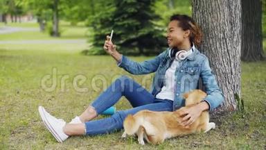 漂亮的年轻女子非裔美国学生正在用智能手机打<strong>视频</strong>电话，并在<strong>草坪</strong>上抚摸她的狗