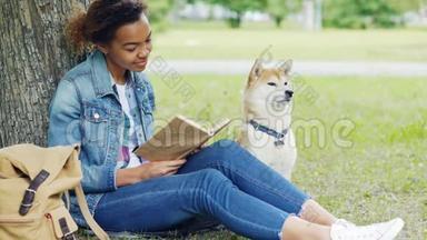 善良的混血女人<strong>正在</strong>公园<strong>看书</strong>，一起抚摸她坐在树下草坪上的狗。 智能智能