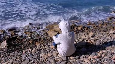 穿着白色羽绒服和风帽的女人坐在岸边，看着汹涌的海浪拍打着石头