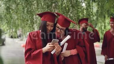 女<strong>毕业生</strong>们用智能手机看屏幕，有说有笑，站在户外拿着文凭，女孩们