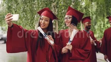 漂亮的女毕业生们正在用智能手机用毕业证书卷轴自拍，女人们在摆姿势和微笑