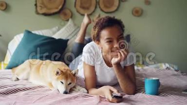 美国黑人妇女正在看电视，手里拿着<strong>遥控</strong>器，按着<strong>按钮</strong>，一边笑着，一边躺着她的宠物狗
