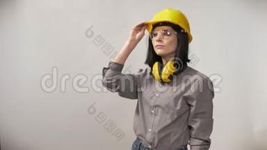 年轻建筑工人妇女戴着耳机、眼镜和头盔，戴着头盔，表情严肃，背景白色