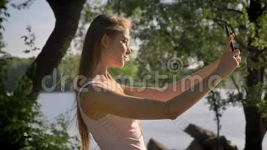 年轻漂亮的女人拿着手机自拍，对着镜头微笑，站在河边的公园里