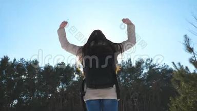 年轻女孩带着背包跳跃，在背景下与冬天的森林一起快乐。 女人举手享受自由