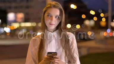 一个年轻漂亮、爱做梦的女孩正在夏天晚上在市中心的智能手机上输入信息