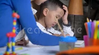 亚洲年轻的母亲给儿子解释，帮孩子做作业，坐在桌子后面，亚洲男孩抱着他的头，看书