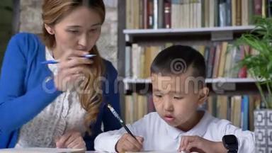 亚洲年轻的母亲和儿子一起写作，女人帮助孩子，坐在桌子后面，亚洲孩子做家庭作业，书架