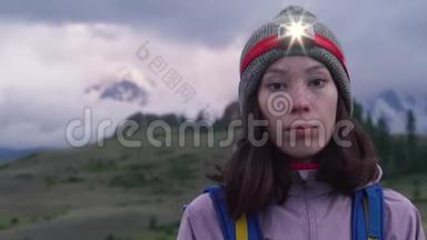 一个年轻的女人带着背包和前照灯在攀爬前看着相机。 背景中的雾山..