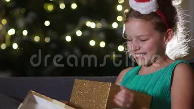 可爱的女孩为圣诞礼物而高兴。 女生正坐在新年树对面的沙发上抱着一个盒子