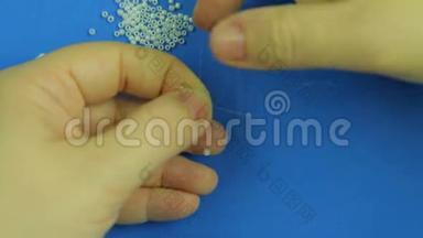 女人`手在针上串着白色的珠子，在蓝色的背景下编织。 特写镜头。 手工制作。