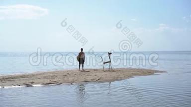 一个带着冲浪板的年轻人站在一只木头<strong>动物</strong>旁边，它是用棍子在<strong>水中</strong>央的一个沙岛上做的。