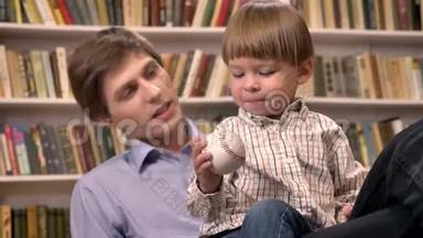 小男孩和他年轻的父亲坐在一起，拿着球，书架上有书背景