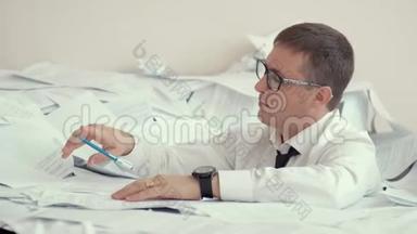 一个戴眼镜的疲惫的年轻人坐在一堆文件和签名文件里。 他胳膊上戴着手表，他累了，