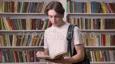 年轻的美男子穿着白衬衫，站在图书馆看书，看着相机，拿着书，书架