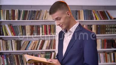 年轻英俊的商人正在<strong>看书</strong>，看相机，微笑，背景<strong>书架</strong>，侧视