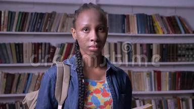 年轻的非裔美国妇女，带着背包在图书馆看书，看着相机，严肃而体贴