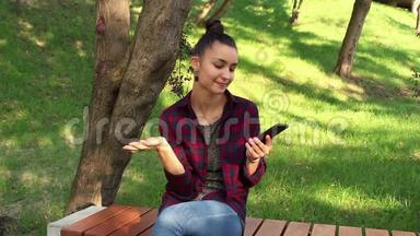 年轻漂亮的女孩坐在公园的长凳上，在手机里浏览社交网络磁带。
