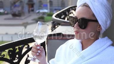优雅的女孩穿着白色的浴袍，头上有一条白色的毛巾，享受着一杯白酒