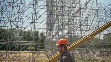年轻建筑工人戴着安全帽，扛着重金属木板，在稻草场上巨大的装置附近工作